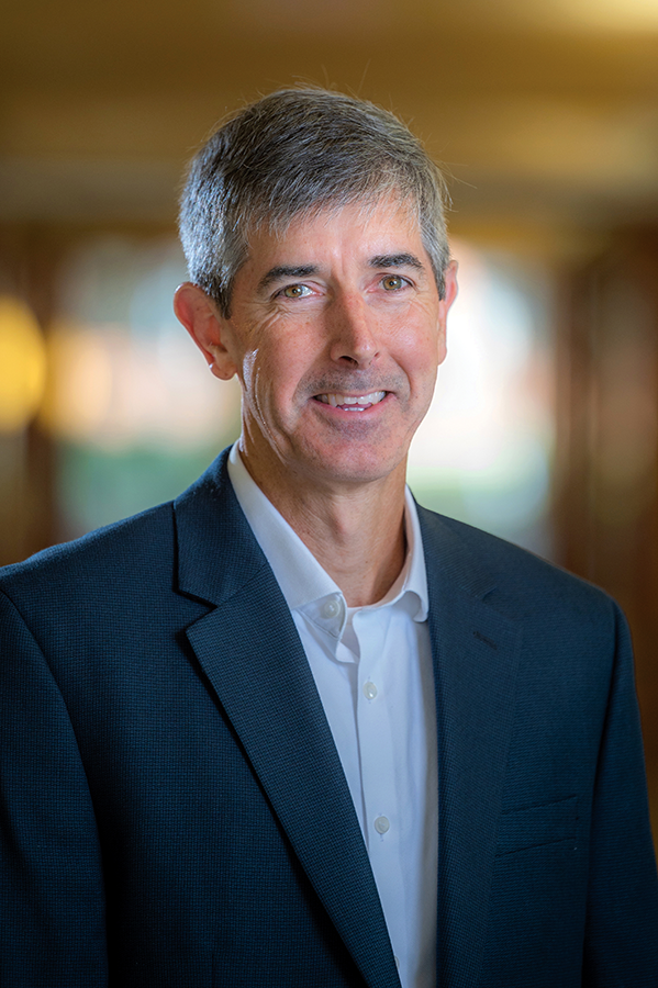 A man in a suit coat is standing in a long university hallway.